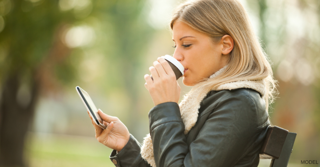 A young woman sitting on a park bench, scrolling her phone and drinking coffee (model)