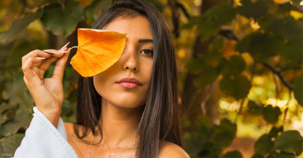 Woman with clear, beautiful skin (model) standing outdoors holding a fallen orange leaf over her left eye.