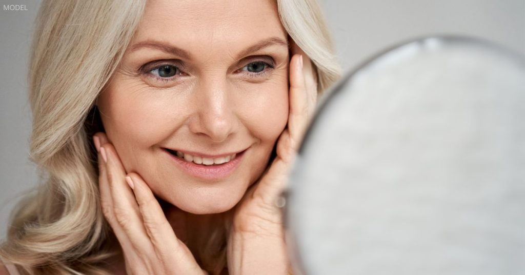 Middle-aged woman eith clear skin (model) looking in the mirror and touching her face.