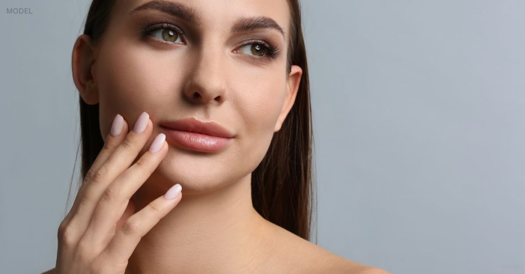 Woman with a slim face and youthful complexion (model) touches a hand to her face.