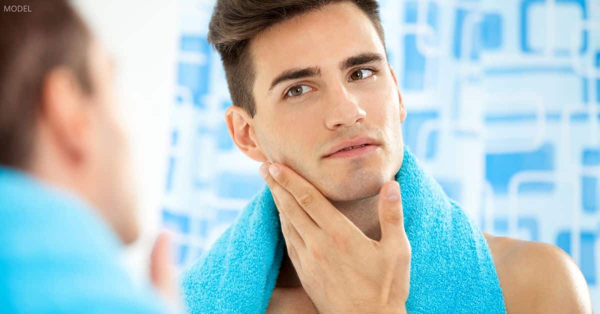 Man with a defined and strong chin (model) places a hand underneath his face while wearing a towel.