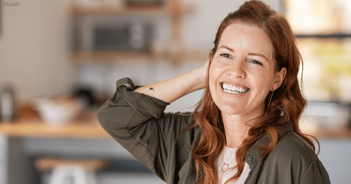 A mature, red-haired woman touching her hair while standing in a kitchen