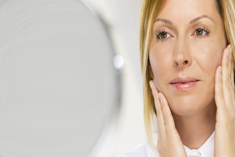 Middle aged woman admires her voluminous cheeks in the mirror after fat transfer