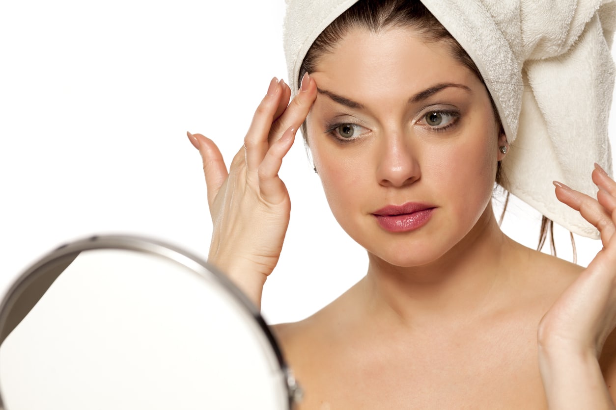 Woman in a towel looking at herself in the mirror and poking at her eyebrow, in need of a brow lift or eyelid surgery.