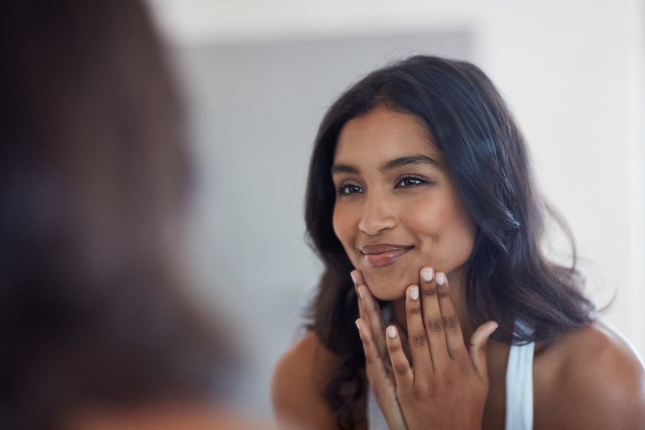 Beautiful young woman smiling and touching her face in the mirror after facial fat transfer for balanced proportions.